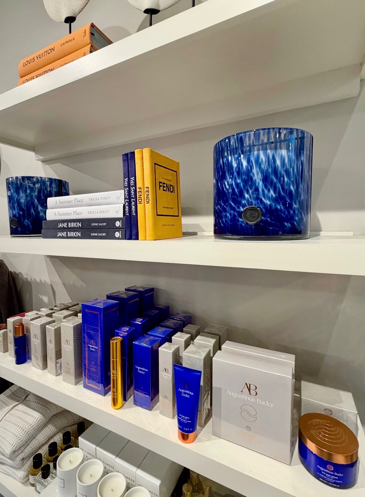 Blue glass vessels, fashion books, fragrance, and skincare products arranged on white Nantucket shelves